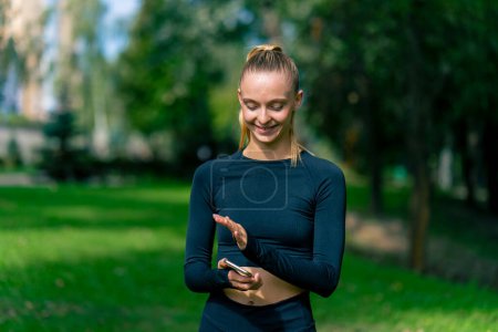 Téléchargez les photos : Une jeune fille en vêtements de sport regarde son écran de téléphone et sourit en faisant du jogging dans le parc le matin - en image libre de droit