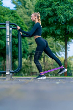 Foto de Atleta chica enfocada realiza ejercicios con una banda elástica deportiva en máquinas de ejercicio callejeras - Imagen libre de derechos