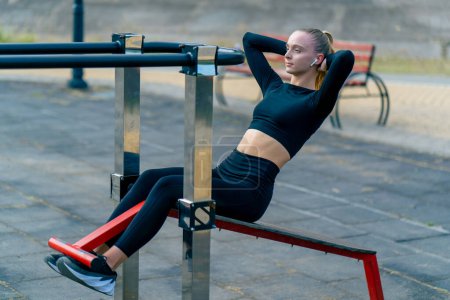 Photo for Athletic young woman pumps up her abs during a morning workout on a sports machine on the street - Royalty Free Image