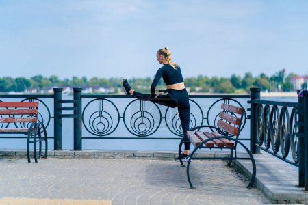 Photo for A girl athlete does morning stretching for leg flexibility on the embankment overlooking the river - Royalty Free Image