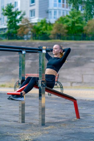 Photo for Athletic young woman pumps up her abs during a morning workout on a sports machine on the street - Royalty Free Image