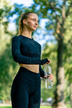 Téléchargez les photos : Jeune femme athlétique après un jogging matinal actif et l'exercice boit de l'eau d'une bouteille sur l'allée dans le parc - en image libre de droit