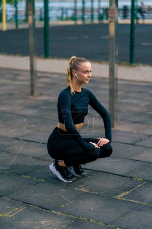 Téléchargez les photos : Belle fille en uniforme de sport effectue squats pour pomper ses jambes sur une aire de jeux de rue sportive - en image libre de droit