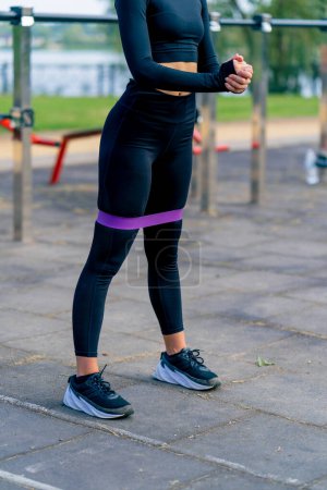 Téléchargez les photos : Jambes d'un entraîneur de sport féminin effectuant des exercices avec une bande élastique pour renforcer les muscles sur le terrain de sport - en image libre de droit