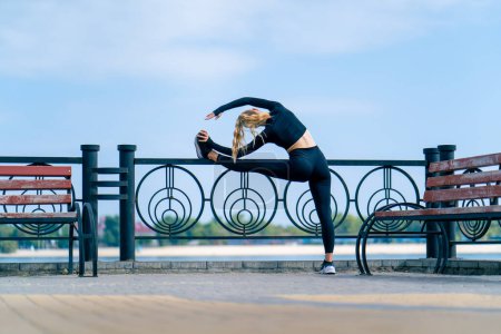 Téléchargez les photos : Une athlète fille fait matin étirement pour la flexibilité des jambes sur le remblai surplombant la rivière - en image libre de droit