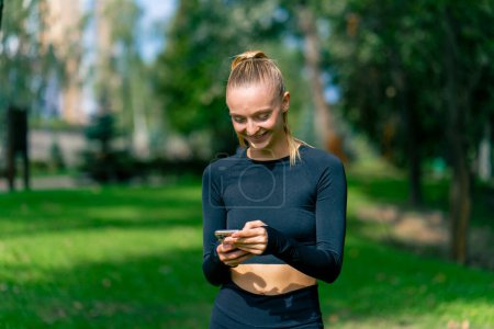 Foto de Una joven en ropa deportiva mira la pantalla de su teléfono y sonríe mientras trota en el parque por la mañana - Imagen libre de derechos