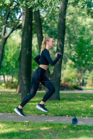 Photo for A young girl in sportswear and headphones runs in the park in the morning to maintain physical fitness - Royalty Free Image