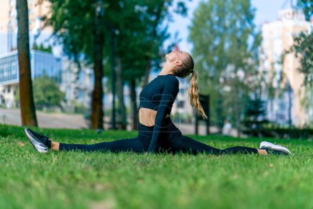 Photo for A young beautiful girl sits on the splits and does stretching after training in the morning in the park - Royalty Free Image