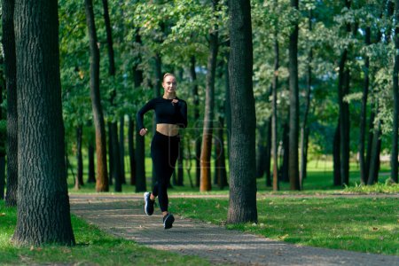 Photo for A young girl in sportswear and headphones runs in the park in the morning to maintain physical fitness - Royalty Free Image