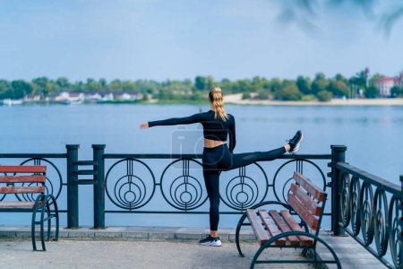 Photo for A girl athlete does morning stretching for leg flexibility on the embankment overlooking the river - Royalty Free Image