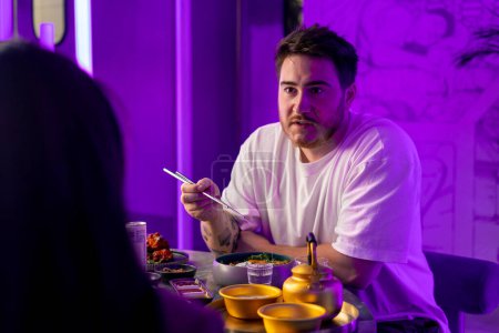 Photo for Portrait of a guy enjoying traditional Korean cuisine with chopsticks in a restaurant and arguing with a girl on date - Royalty Free Image