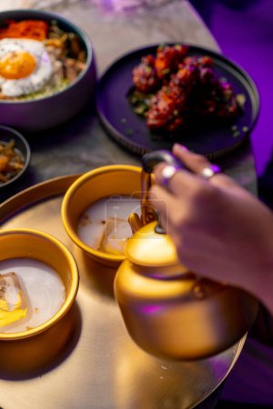 Téléchargez les photos : Gros plan d'une fille versant un makoli traditionnel coréen à faible teneur en alcool d'une théière dorée dans une tasse avec de la glace au restaurant - en image libre de droit