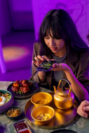 Foto de Una joven toma una foto en un teléfono inteligente de servir una bebida en una tetera en un restaurante coreano - Imagen libre de derechos