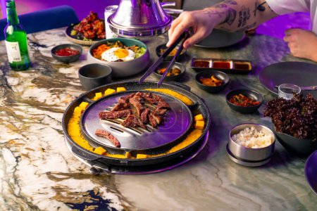 Téléchargez les photos : Gros plan d'un couple amoureux à une table déjeunant dans un restaurant coréen cueillant de la viande juteuse du gril avec des pinces - en image libre de droit