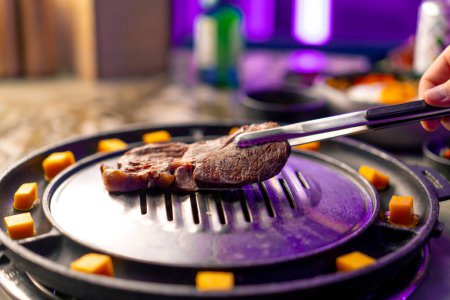 Téléchargez les photos : Gros plan d'une main avec pinces ramasser un morceau de viande cru juteux à mettre sur le gril dans le restaurant coréen - en image libre de droit