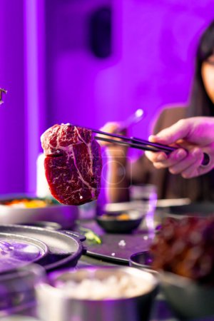 Photo for Close-up of a hand with tongs picking up a juicy raw piece of meat to put on the grill in Korean restaurant - Royalty Free Image