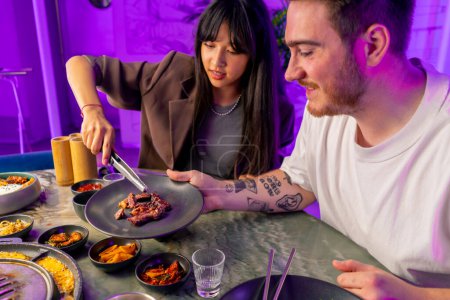 Foto de Una pareja enamorada cenando en un restaurante coreano tomar carne jugosa de la parrilla con pinzas comer con palillos - Imagen libre de derechos