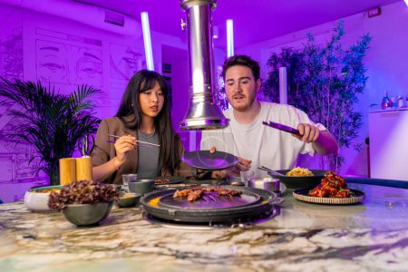 Photo for A couple in love having dinner in a Korean restaurant take juicy meat from the grill with tongs eat with chopsticks - Royalty Free Image