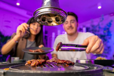 Photo for A couple in love having dinner in a Korean restaurant take juicy meat from the grill with tongs eat with chopsticks - Royalty Free Image