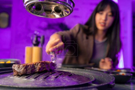 Foto de Una chica voltea un jugoso pedazo de carne a la parrilla con pinzas en un restaurante coreano para freír tratar a un amigo - Imagen libre de derechos