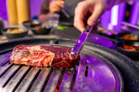 Téléchargez les photos : Gros plan d'une main avec pinces ramasser un morceau de viande cru juteux à mettre sur le gril dans le restaurant coréen - en image libre de droit