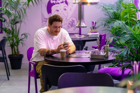 Téléchargez les photos : Un jeune homme est assis à une table dans un café restaurant dans un bel intérieur attendant sa commande avec smartphone à la main - en image libre de droit
