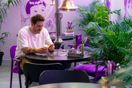 Foto de Un joven está sentado en una mesa en un restaurante cafetería en un hermoso interior esperando su orden con un teléfono inteligente en la mano - Imagen libre de derechos
