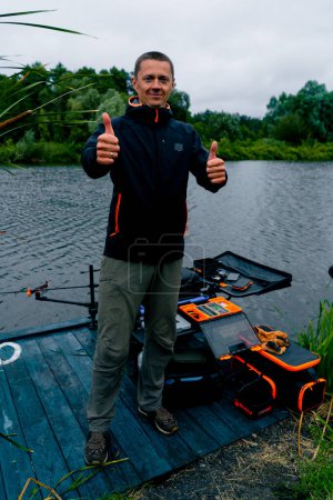 Foto de Retrato de un pescador sonriente con una caña de pescar o una caña de hilar y herramientas profesionales muestra una señal estupenda con su mano en la pesca del río - Imagen libre de derechos