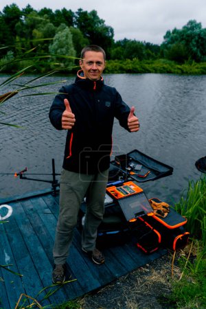 Téléchargez les photos : Portrait d'un pêcheur souriant avec une canne à pêche ou une canne à pêche et des outils professionnels montre un super panneau avec sa main sur la pêche fluviale - en image libre de droit