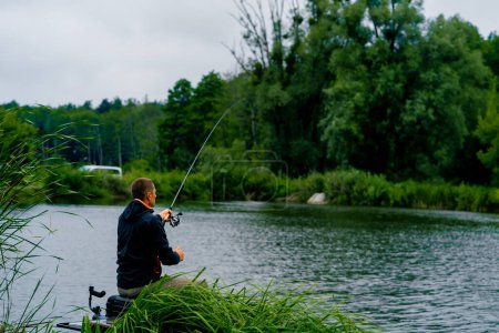 Foto de Pescador con caña de pescar o spinning y herramientas profesionales sentado en la orilla del río vista trasera Tire de peces de la pesca deportiva lago - Imagen libre de derechos
