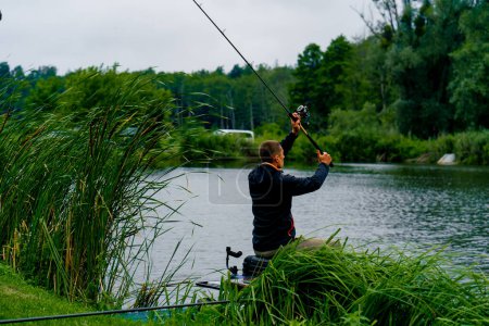 Téléchargez les photos : Pêcheur avec canne à pêche ou filature et outils professionnels assis sur la rive de la rivière vue arrière Sortir le poisson de la pêche sportive du lac - en image libre de droit