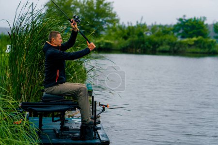 Téléchargez les photos : Pêcheur jette une canne à pêche ou une canne à pêche dans l'eau tout en étant assis sur la rive de la rivière sur la jetée Tirez le poisson hors du lac pêche sportive - en image libre de droit