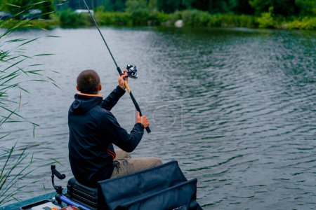 Foto de Pescador con caña de pescar o spinning y herramientas profesionales sentado en la orilla del río vista trasera Tire de peces de la pesca deportiva lago - Imagen libre de derechos