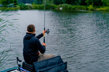 Téléchargez les photos : Pêcheur avec canne à pêche ou filature et outils professionnels assis sur la rive de la rivière vue arrière Sortir le poisson de la pêche sportive du lac - en image libre de droit