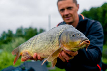 Photo for Portrait of a satisfied professional fisherman holding a carp on the bank of a river fishing in a pond with good catch - Royalty Free Image