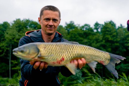 Photo for Portrait of a satisfied professional fisherman holding a carp on the bank of a river fishing in a pond with good catch - Royalty Free Image