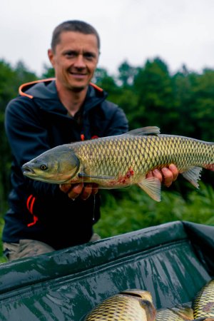 Téléchargez les photos : Portrait d'un pêcheur professionnel satisfait tenant une carpe sur le bord d'une rivière pêchant dans un étang avec de bonnes prises - en image libre de droit