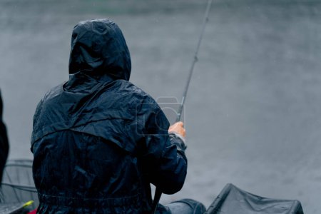 Foto de Pescador en impermeable con caña de pescar o herramientas profesionales de spinning sentado en la orilla del río vista trasera Tire de los peces de la pesca deportiva lago - Imagen libre de derechos