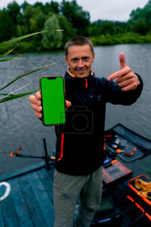 Foto de Retrato de un pescador sonriente con herramientas de spinning y profesionales sosteniendo un smartphone con una pantalla verde en la orilla del río pesca deportiva - Imagen libre de derechos