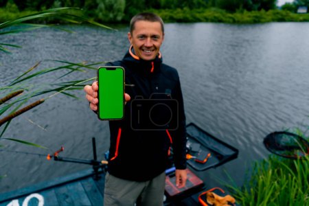 Photo for Portrait of a smiling fisherman with spinning and professional tools holding a smartphone with a green screen on river bank sport fishing - Royalty Free Image