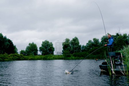 Téléchargez les photos : Pêcheur avec canne à pêche ou filature outils professionnels debout sur le bord de la rivière Sortir le poisson du lac à l'aide d'un filet de pêche sportive - en image libre de droit
