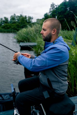 Téléchargez les photos : Pêcheur avec canne à pêche ou canne à pêche et outils professionnels assis sur le bord de la rivière Sortir le poisson de la pêche sportive du lac - en image libre de droit