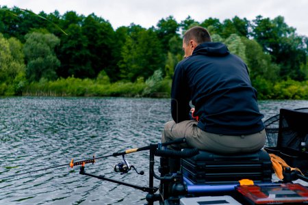 Photo for Fisherman with fishing rod or spinning and professional tools sitting on the river bank rear view Pull fish out of lake sports fishing - Royalty Free Image