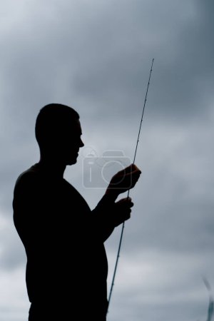 Téléchargez les photos : Silhouette sombre d'un pêcheur avec une canne à pêche sur étang sport ou pêche amateur - en image libre de droit
