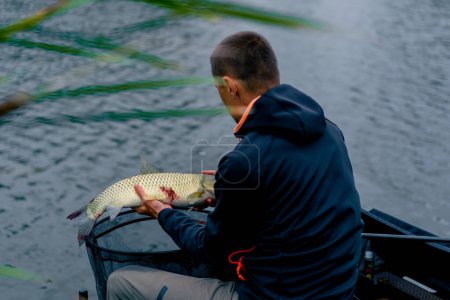 Téléchargez les photos : Portrait d'un pêcheur professionnel tenant un poisson de carpe sur la rive de la pêche fluviale dans des réservoirs une bonne prise - en image libre de droit