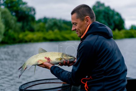 Téléchargez les photos : Portrait d'un pêcheur professionnel tenant un poisson de carpe sur la rive de la pêche fluviale dans des réservoirs une bonne prise - en image libre de droit