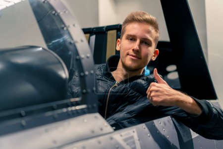 Téléchargez les photos : Satisfait jeune homme assis dans le simulateur de vol de l'avion militaire après vol virtuel montre signe de main super amusant - en image libre de droit