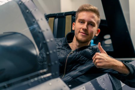 Téléchargez les photos : Satisfait jeune homme assis dans le simulateur de vol de l'avion militaire après vol virtuel montre signe de main super amusant - en image libre de droit