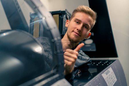 Photo for Satisfied young guy sitting in flight simulator of military plane after virtual flight shows hand sign super fun - Royalty Free Image