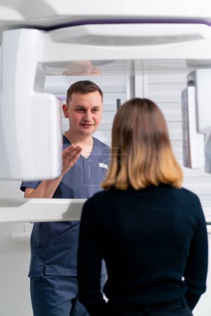 Foto de Un médico masculino consulta a una paciente femenina cerca del dispositivo de exploración 3D antes del inicio del procedimiento en una clínica de cirugía plástica o odontología - Imagen libre de derechos